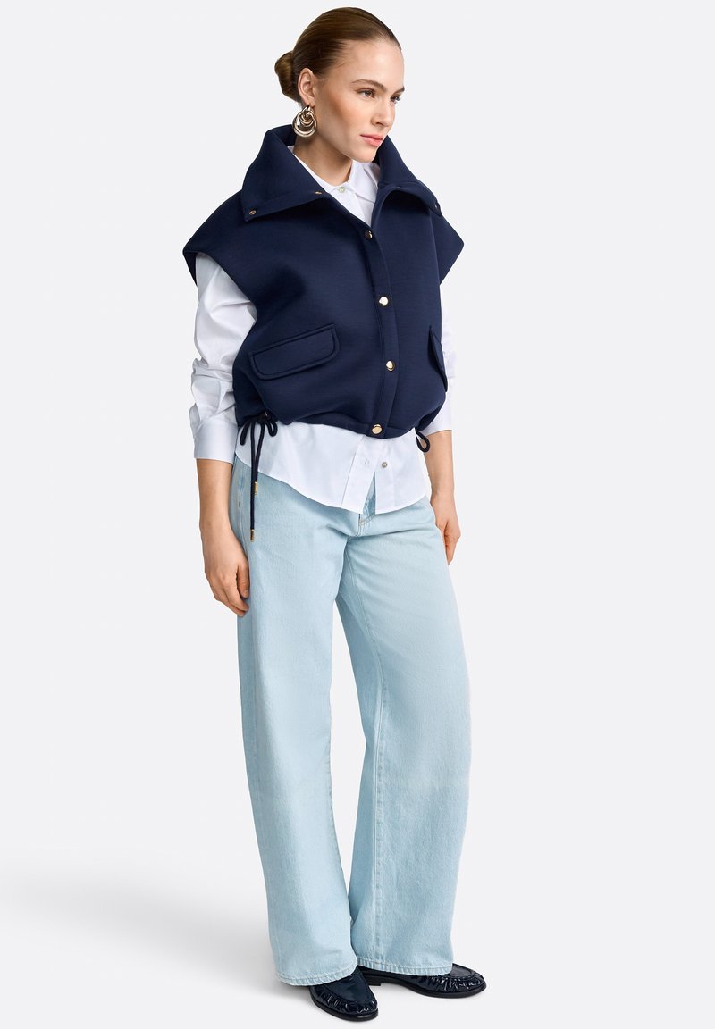 Žena oblečená v námorníckej modrej vesti bez rukávov cez biele košeľové tričko, svetlomodré široké džínsy, čierne topánky a veľké zlaté náušnice.