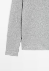Partie d'une chemise manches longues unie gris clair montrant la manche et la partie basse du torse sur fond blanc.