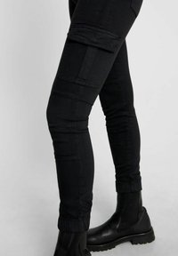 Pantalones cargo negros con diseño ajustado, múltiples bolsillos en los muslos, tobillos ceñidos y una textura suave, combinados con botas negras hasta el tobillo.