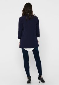 Blazer marine à manches longues, au design ajusté, associé à une chemise blanche et un jean foncé, complété par des chaussures à talons hauts noires.