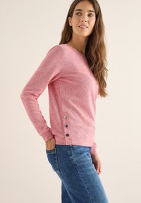 Sudadera de punto rosa jaspeado con mangas largas, cuello redondeado y tres botones decorativos en la costura del lado izquierdo. Combinada con jeans de mezclilla.