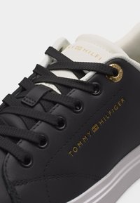 Gros plan d’une basket noire et blanche avec des lacets noirs et le logo Tommy Hilfiger doré sur le côté et la languette.