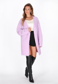 Lila oversized cardigan, jossa on pitkät hihat, avonainen etuosa ja pörröinen pinta, yhdistettynä vaaleanvihreään toppiin ja mustaan minihameeseen, jota korostavat korkeat mustat saappaat.