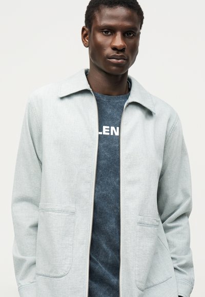 Νεαρός άνδρας που φοράει ανοιχτό γκρι ζακέτα με φερμουάρ πάνω από μπλε σκούρο πουκάμισο με μερικώς ορατό λευκό κείμενο, στέκεται μπροστά σε απλό φόντο.