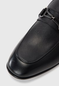 Chaussure en cuir noir avec une surface texturée, un bout pointu et une sangle décorative avec boucle. Fini lisse avec des accents contrastés.