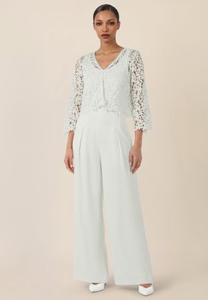 Woman wearing white lace three-quarter sleeve top with V-neck and high-waist wide-leg white pants, standing against beige background.