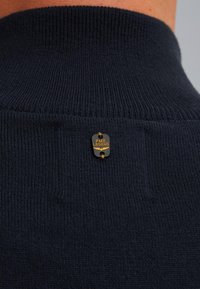 Tamno plavi rebrasti pleteni materijal s malom crnom i zlatnom oznakom brenda PME Legend koja je ušivena blizu ovratnika.