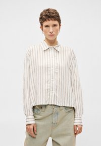 Gaismas krēmkrāsas audumā, ar vertikālām bežām svītrām, ar pogām aizpogājama garām piedurknēm. Iezīmēta ar norādītu apkakli un platām, brīvi pieguļošām piedurknēm.