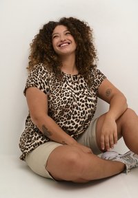 T-shirt à manches courtes en imprimé léopard beige et noir, associé à un short beige clair. Cheveux bouclés ; portant des baskets avec des détails texturés.