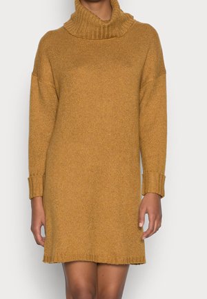 Kvinde iført en sennepsgul strikket sweaterkjole med en chunky høj krave og lange ærmer, stående foran en ensartet lys baggrund.