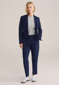 Jūras zila bleizeris un bikses ar pelēku džemperi zem tā. Sneakeri ar baltām un melnām akcentiem. Gluda auduma un pielāgota piegriezuma.