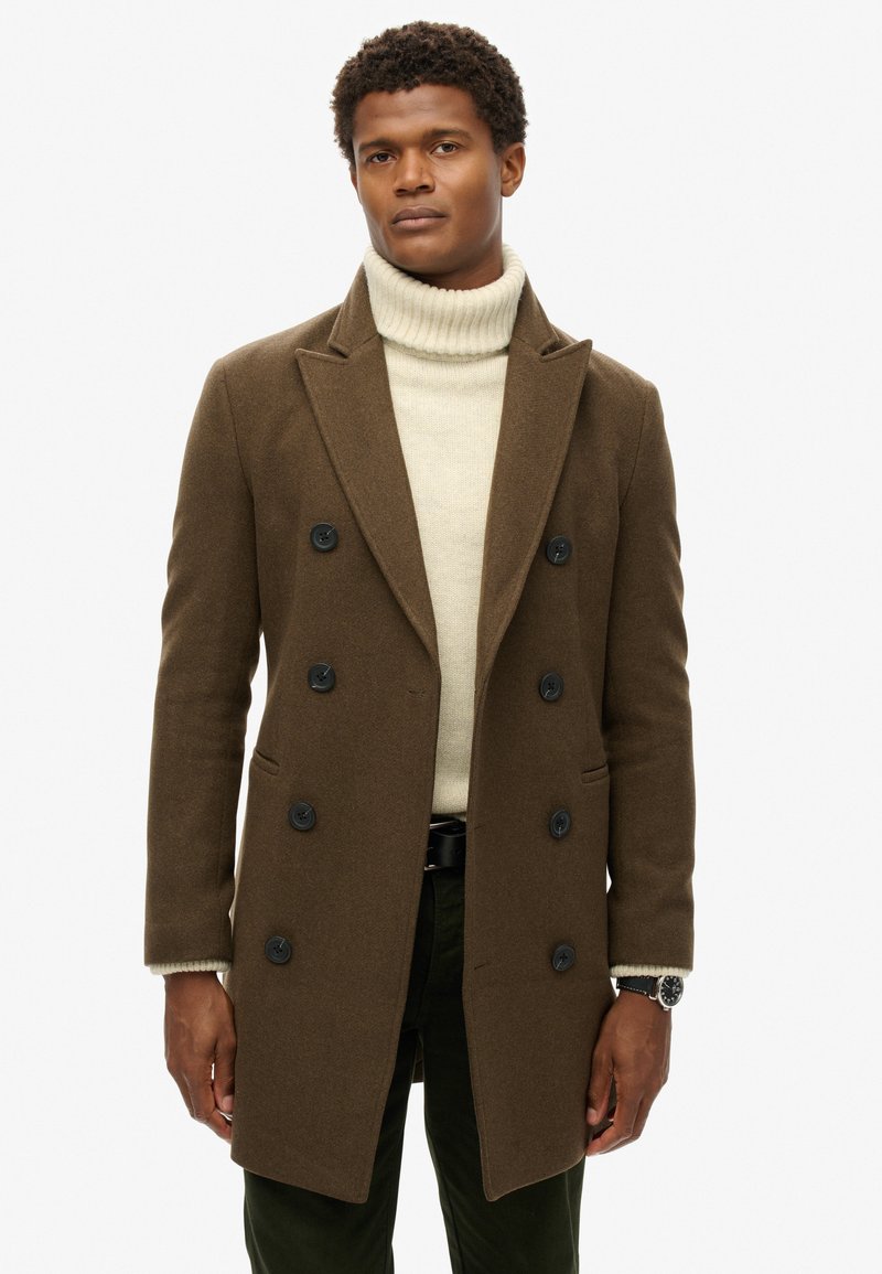 Brun uld dobbeltbrystet frakke med slåede kraver, sorte knapper, sidelommer og en beige rullekrave sweater under.