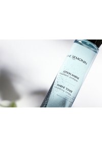 Heldere glazen fles met blauwe vloeistof en belletjes. Etiket bevat "Lotion Marine" en "Marine Toner" in zwart tekst. Zwarte deksel.