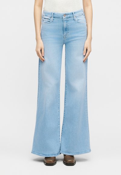 Άτομο που φοράει ανοιχτό μπλε τζιν με ψηλή μέση και φαρδύ πόδι, καθώς και καφέ παπούτσια, στέκεται μπροστά σε ένα απλό λευκό φόντο.