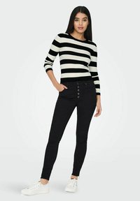 Zwarte skinny jeans met een hoge taille en knopen aan de voorkant, gecombineerd met een zwart-wit gestreept ribbeltrui. Witte sneakers.