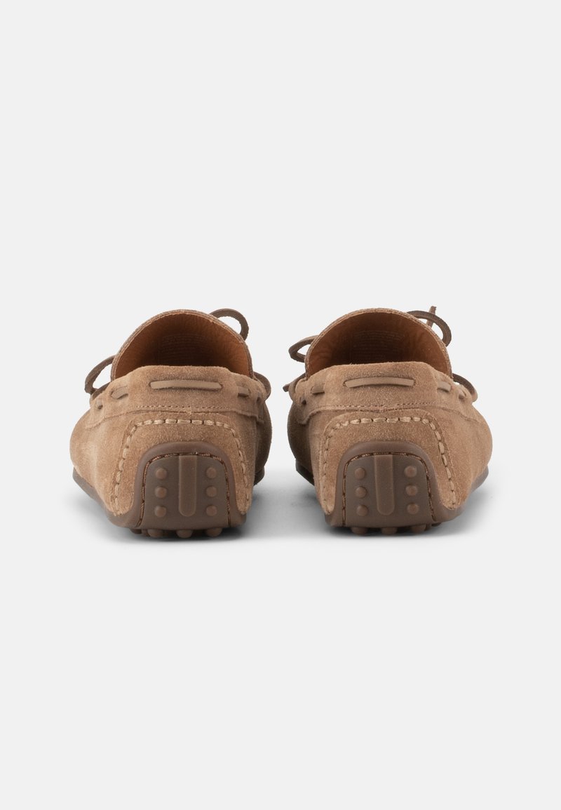 Een paar bruine suède mocassin loafers bekeken vanaf de achterkant, met rubberen nopjes op de hak voor grip, geplaatst op een witte achtergrond.