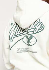 Felpa crema realizzata in misto cotone, con grande testo verde e una grafica di un'aquila sul retro. Texture morbida con un cappuccio ampio.