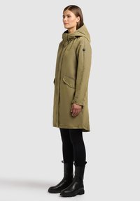 Vrouw draagt een knie-lange olijfgroene gewatteerde jas met capuchon, zwarte legging en zwarte laarzen tot halverwege het been, staand op de zij op een lichtgrijze achtergrond.