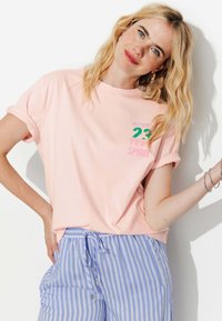 T-shirt rose clair avec des manches retroussées, arborant un texte vert et rose "AUTHENTIC 23 TEAM SPIRIT". Associé à un pantalon rayé bleu.