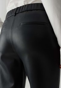 Pantalons en simili cuir noir avec une texture lisse, taille élastique et deux poches arrière, présentés en contre-plongée.