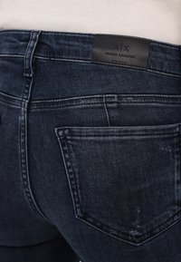 Jean en denim bleu foncé avec une poche arrière et une étiquette en cuir noir Armani Exchange sur la ceinture.