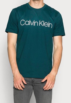 Homme portant un t-shirt vert foncé à manches courtes avec un logo Calvin Klein blanc et un jean gris sur un fond clair uni.