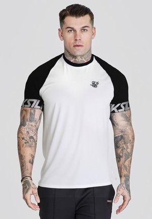 Mann mit Tätowierungen trägt ein weißes und schwarzes kurzärmliges Hemd mit Markenärmeln und steht vor einem schlichten hellgrauen Hintergrund.