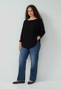 Zwarte, oversized gebreide top met een brede halslijn, gecombineerd met blauwe straight-leg jeans. De schoenen hebben een zwart, gestructureerd patroon met een platte vorm.