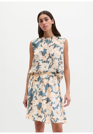 Donna che indossa un vestito senza maniche con motivo floreale astratto blu, beige e nero, in piedi davanti a uno sfondo chiaro e uniforme.