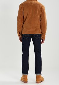 Veste en polaire marron avec col, associée à un jean bleu foncé et des bottes de travail beiges. Le tissu semble doux et texturé.