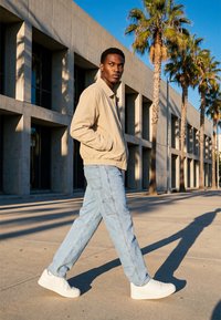 Mann i beige jakke, lyseblå jeans og hvite joggesko går forbi en moderne betongbygg med palmer i sollys.