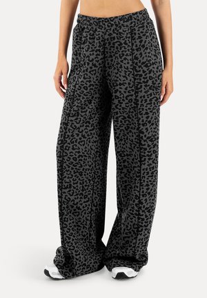 Kobieta ubrana w luźne, szerokie szare spodnie z czarnym nadrukiem w panterkę i białe trampki, stojąca na tle jednolitego tła.