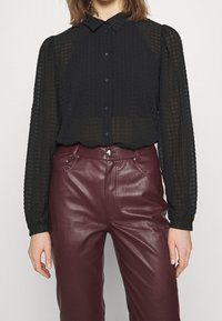 Blouse noire texturée avec de longues manches transparentes et fermeture à boutons, associée à un pantalon en cuir bordeaux taille haute.