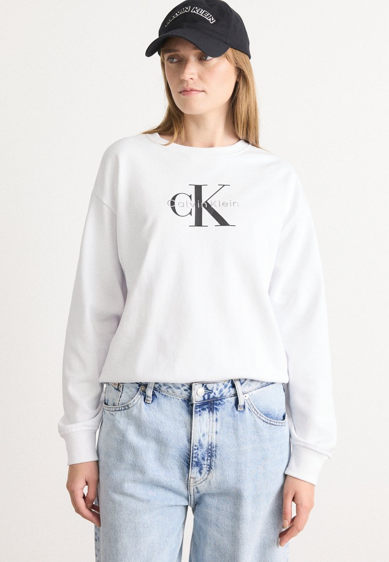 Femme portant un sweatshirt blanc de Calvin Klein, un jean bleu clair et une casquette de baseball noire de Calvin Klein, se tenant devant un fond uni.