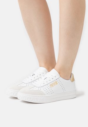 Baskets blanches basses à lacets sur le devant, logo doré "Versace Jeans Couture" sur le côté et le talon, portées pieds nus sur un fond blanc.