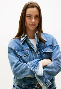 Giubbotto di jeans di colore azzurro chiaro con due tasche sul petto, chiusura con bottoni e camicia interna a contrasto con righe verticali. Il modello ha le braccia incrociate.