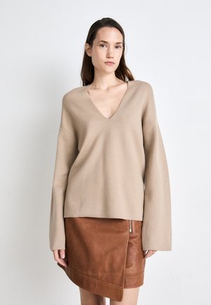 Beige V-Ausschnitt-Pullover mit langen Ärmeln kombiniert mit einem braunen Lederwickelrock, der mit einem seitlichen Reißverschluss versehen ist. Weicher Stoff, lässige Passform.