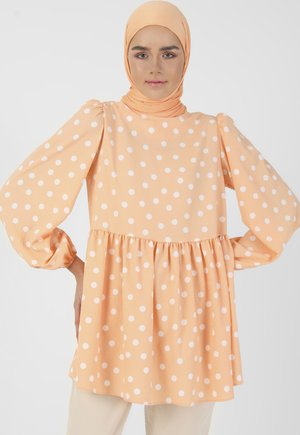 Peach-colored blouse with white polka dots, flared sleeves, gathered waist, and soft texture. Paired with light-colored pants.