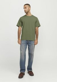 Camiseta verde oliva con cuello redondo, mangas cortas y corte relajado, combinada con jeans claros desgastados y zapatos marrones con cordones.