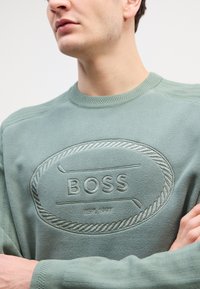 Felpa verde menta in morbido tessuto con logo "BOSS" in rilievo all'interno di un design a corda. Collo e polsini a coste, dettagli di texture minimi.