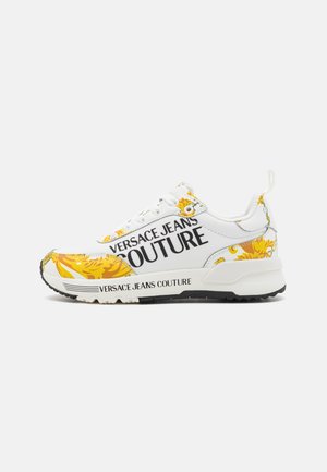 Baskets blanches avec des motifs floraux jaunes, texte "VERSACE JEANS COUTURE", col rembourré et semelle en caoutchouc noir texturé.
