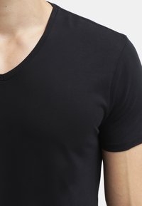 Nærbillede af en person iført en ensfarvet sort V-hals kortærmet T-shirt, der viser halsudskæringen og overarmen mod en lys baggrund.