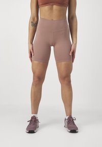 Hoge taille, lichtbruine fietsbroek van rekbare stof. Soepele textuur met gestikte naden. Gecombineerd met lichtpaarse sportschoenen.