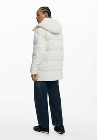 Weißer Pufferjacke mit Kapuze, ausgestattet mit einem gesteppeten Design, sichtbaren Nähten und strukturierten Akzenten. Kombiniert mit dunklen Jeans und schwarzen Schuhen.