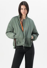 Joven mujer con cabello oscuro lacio lleva una chaqueta bomber verde oliva suelta sobre una blusa naranja y pantalones negros, de pie frente a un fondo blanco.