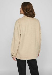 Valkoinen ylisuuri takki, joka on valmistettu pehmeästä villasekoitteesta. Ominaisuuksia ovat kauluksellinen pääntie, pitkät hihat ja napit yksityiskohtina hihoissa.