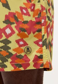 Shorts de bain jaunes avec un motif géométrique multicolore comprenant des teintes d'orange, de rouge, de vert et de blanc. Comprend un patch logo de tête de mort rond.