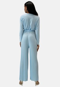 Mono de manga larga azul claro con diseño plisado, que presenta un cuerpo ajustado, pantalones de pierna ancha y una cintura atada. Tejido suave y fluido.