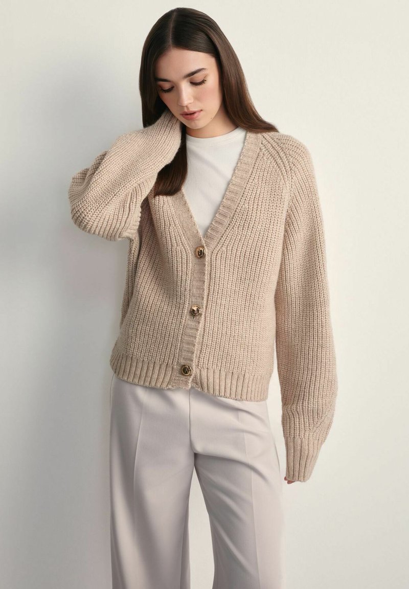 Beżowy sweter dzianinowy z dekoltem w kształcie V, z prążkowanymi mankietami i dołem, oraz trzema złotymi guzikami z przodu. Noszony na jasnej bluzce.
