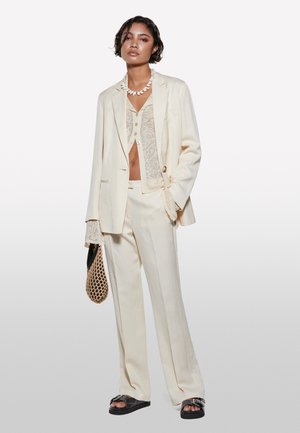 Abito su misura bianco sporco con blazer strutturato e pantaloni a gamba larga, sovrapposto a un top di pizzo. Tessuto testurizzato e design minimalista.